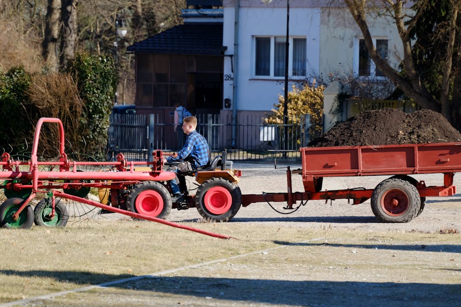 A young boy wearing a blue checkered shirt is seated on a small orange tractor attached to a red trailer filled with dark soil, which is part of outdoor landscape work in a suburban garden. The tracto