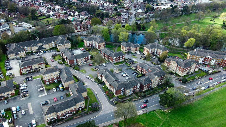 An aerial view of Whitton Park showing a lush green landscape with various trees and shrubs, some with white blossoms, surrounding well-maintained lawns and garden beds. The park layout includes windi